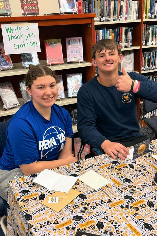 Two smiling students sit at a table with a tablecloth.
