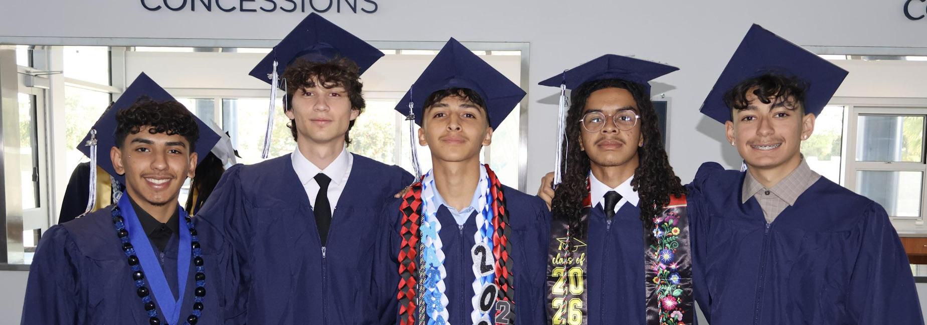 Five male graduates in navy gowns, smiling together with various honors cords.