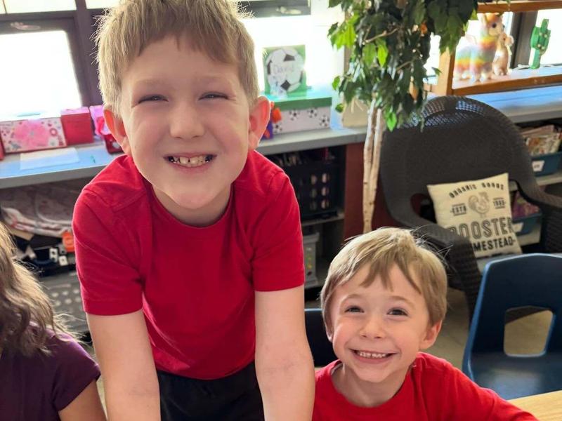 Two kids smiling while coloring at a classroom table.