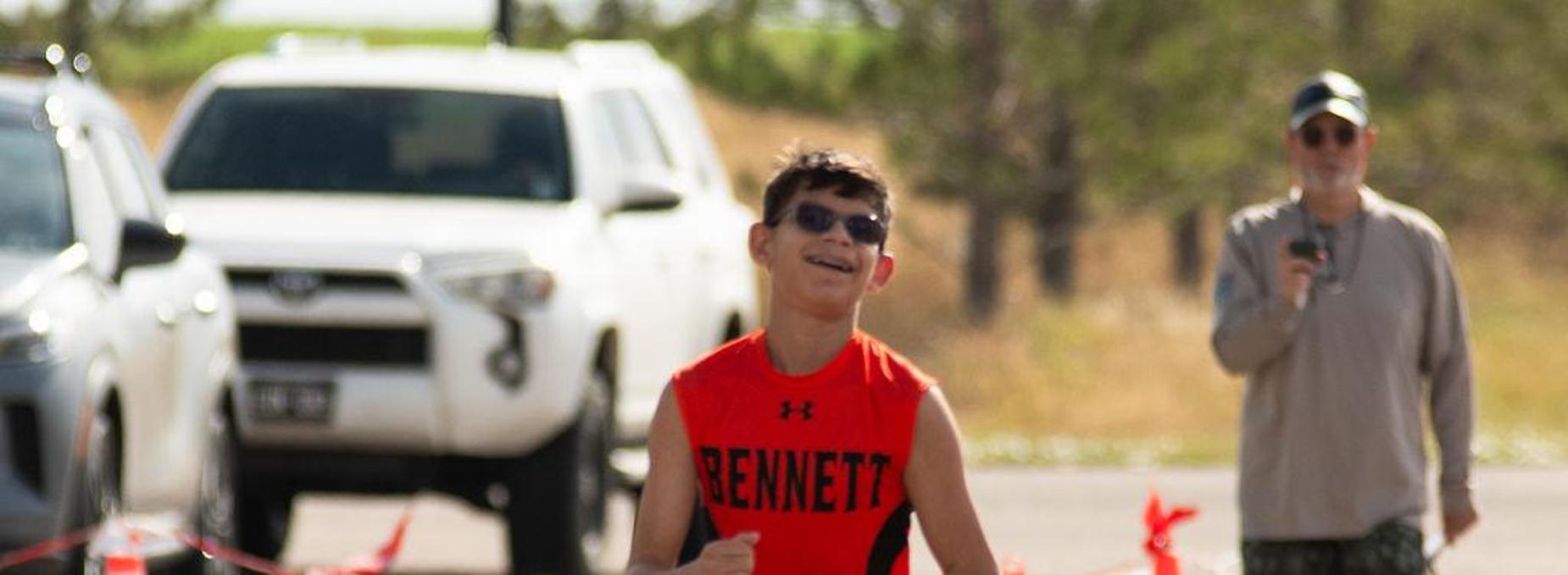 A young boy in a red athletic shirt is running, smiling, near traffic cones and spectators.