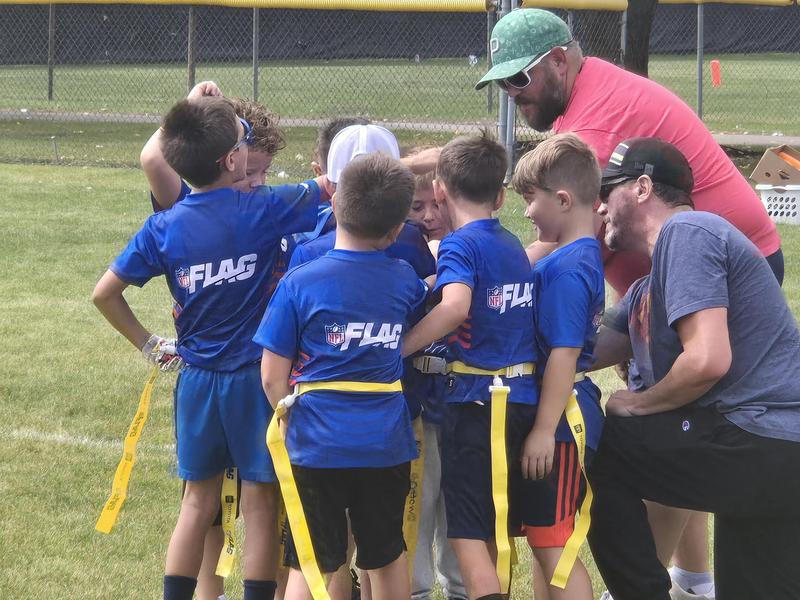 Children in blue jerseys huddle together on a grassy field with a coach for Flag Football.