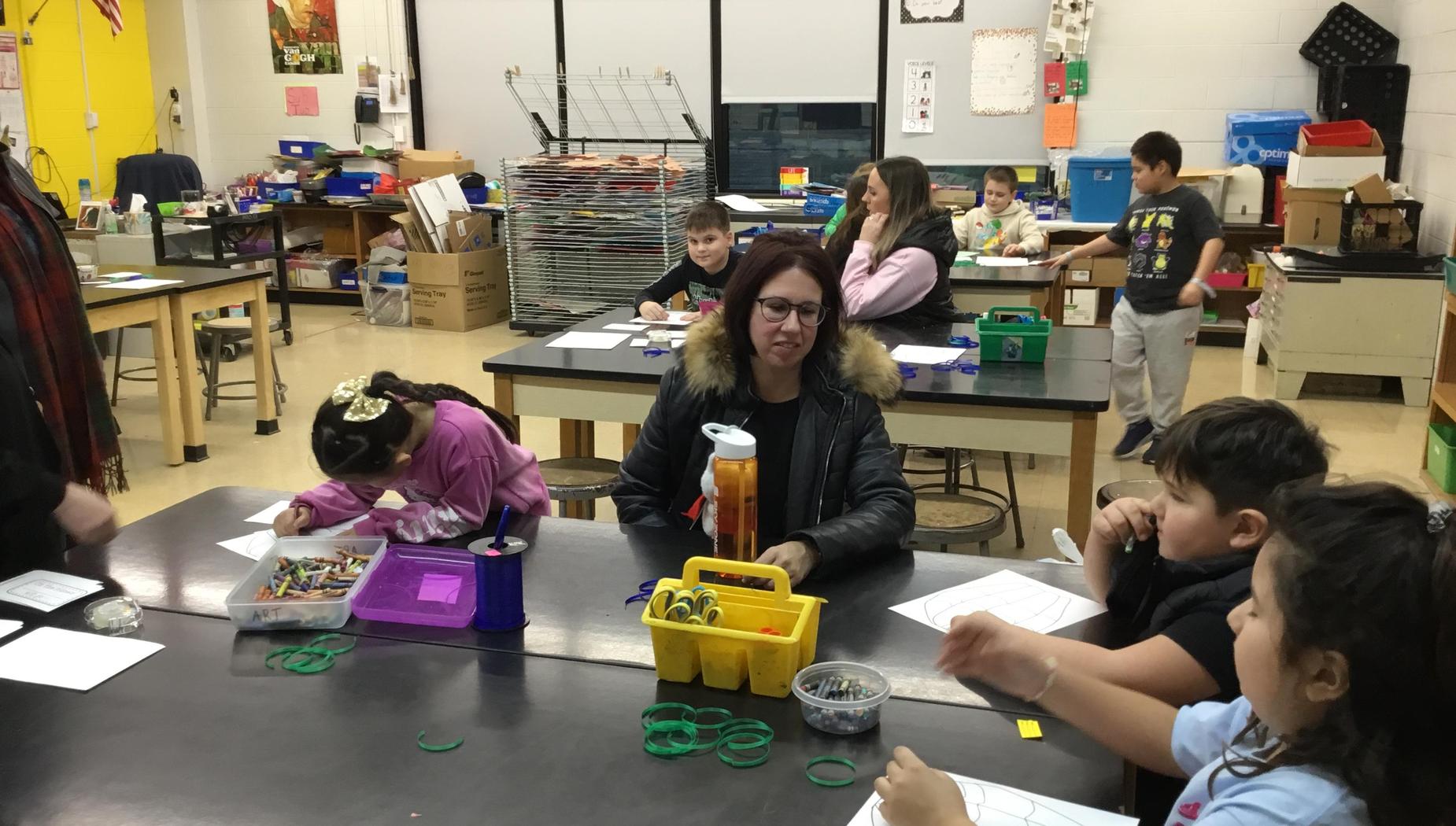 Students and adults participate in activities at a classroom with tables and supplies.