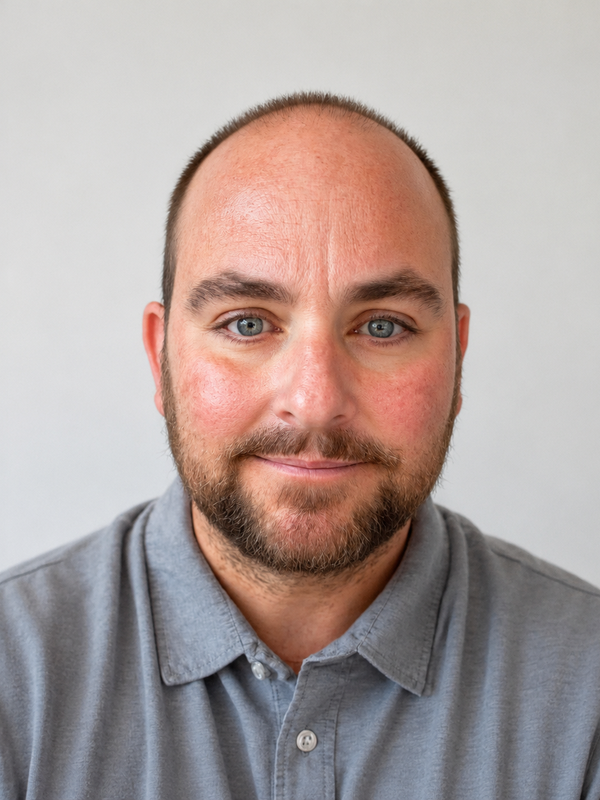 A man with a beard and short hair, wearing a grey shirt, smiling softly.