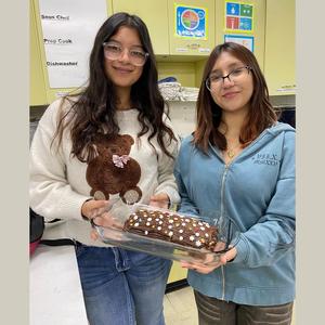 Two students display a chocolate dessert with decorations in a kitchen.