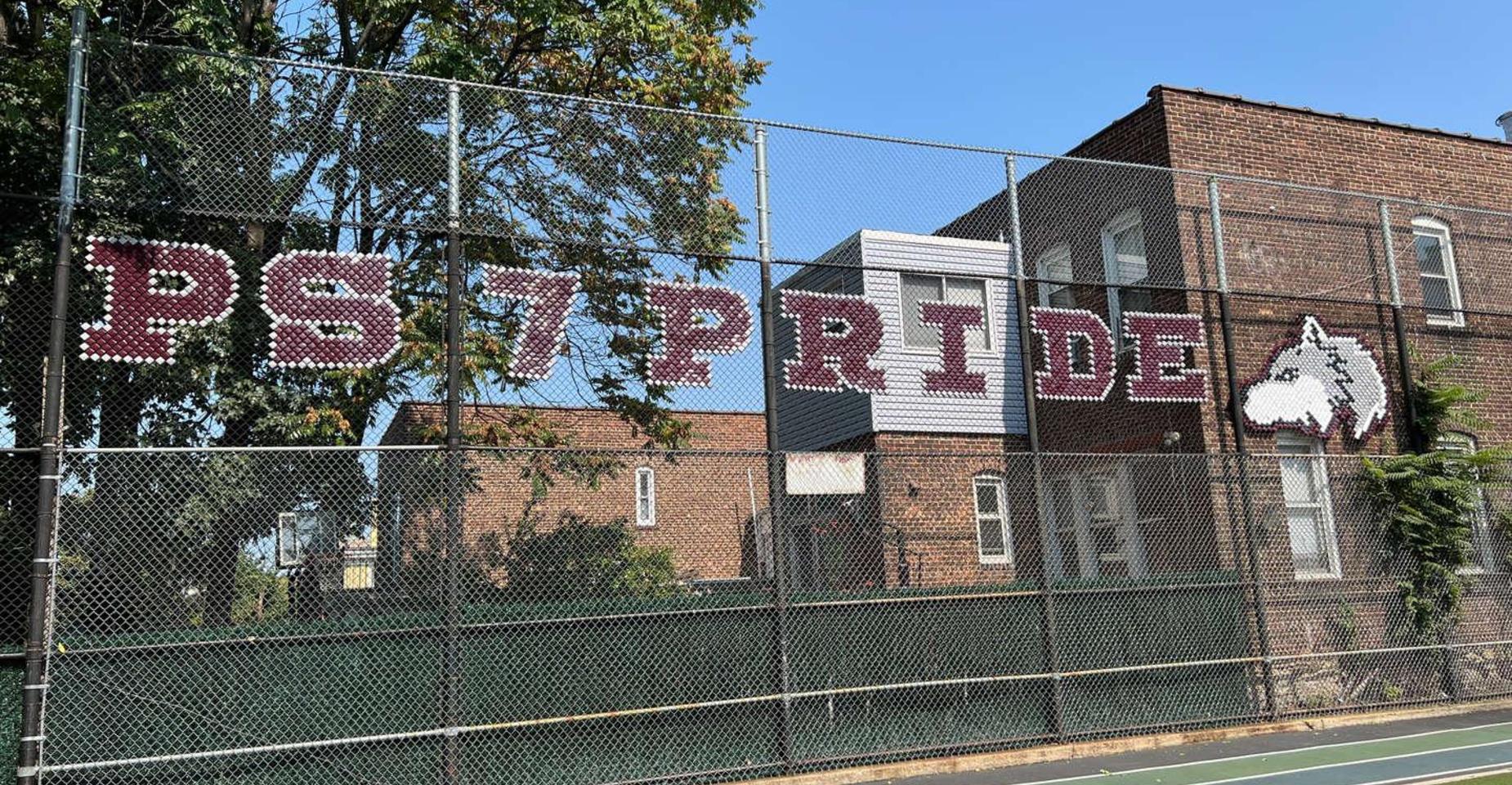 Text reading 'PS 7 PRIDE' on a fence near a brick building.