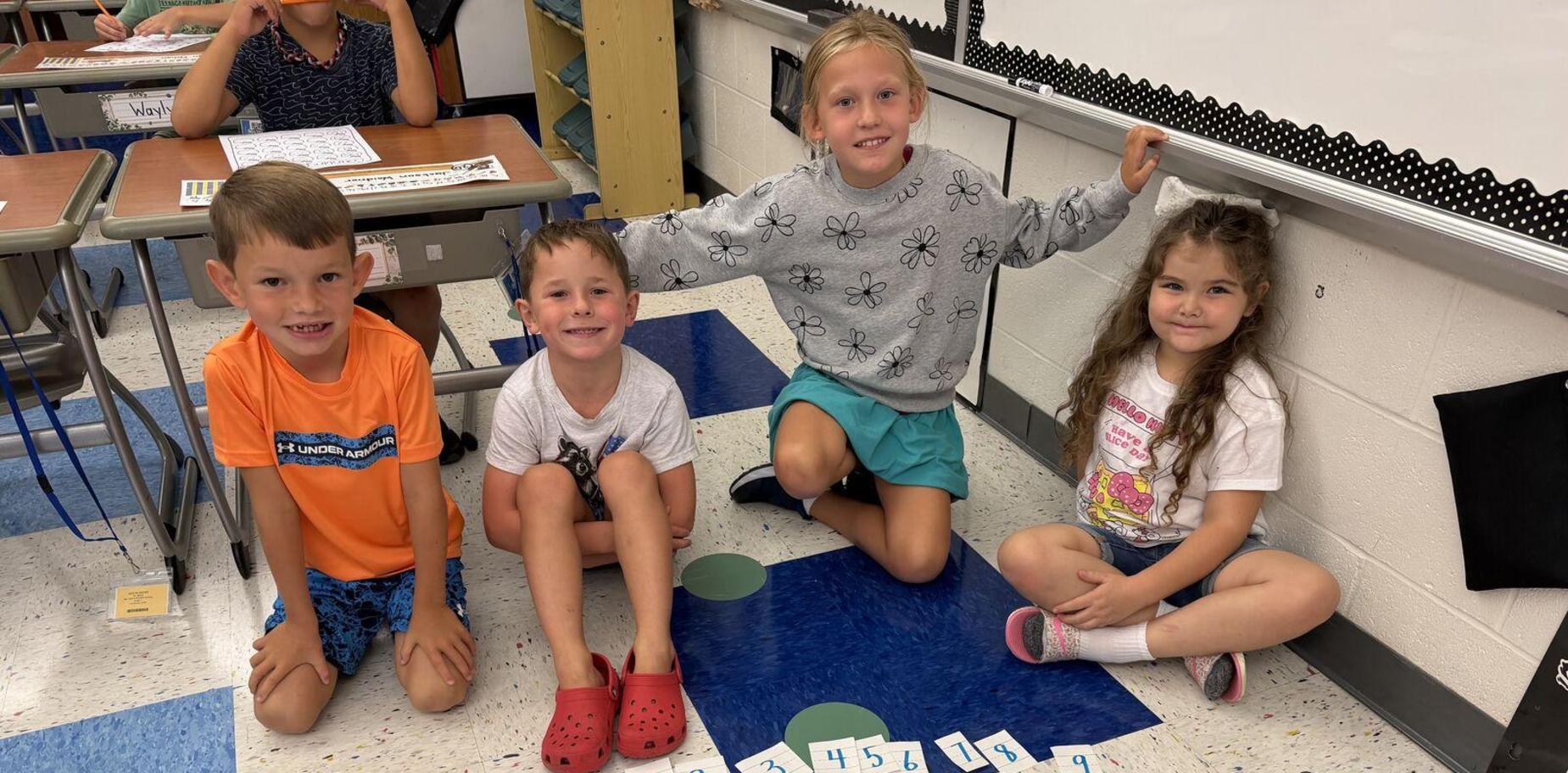 Four children sitting on the floor with number cards in a classroom.