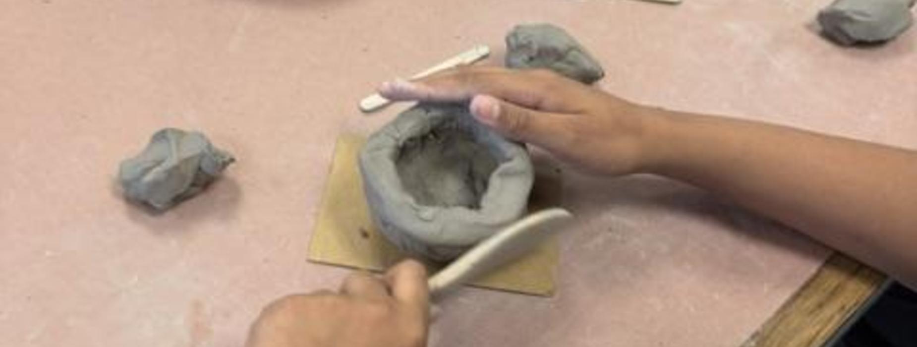 A child's hands shape clay into a pot on a workspace covered with clay tools.