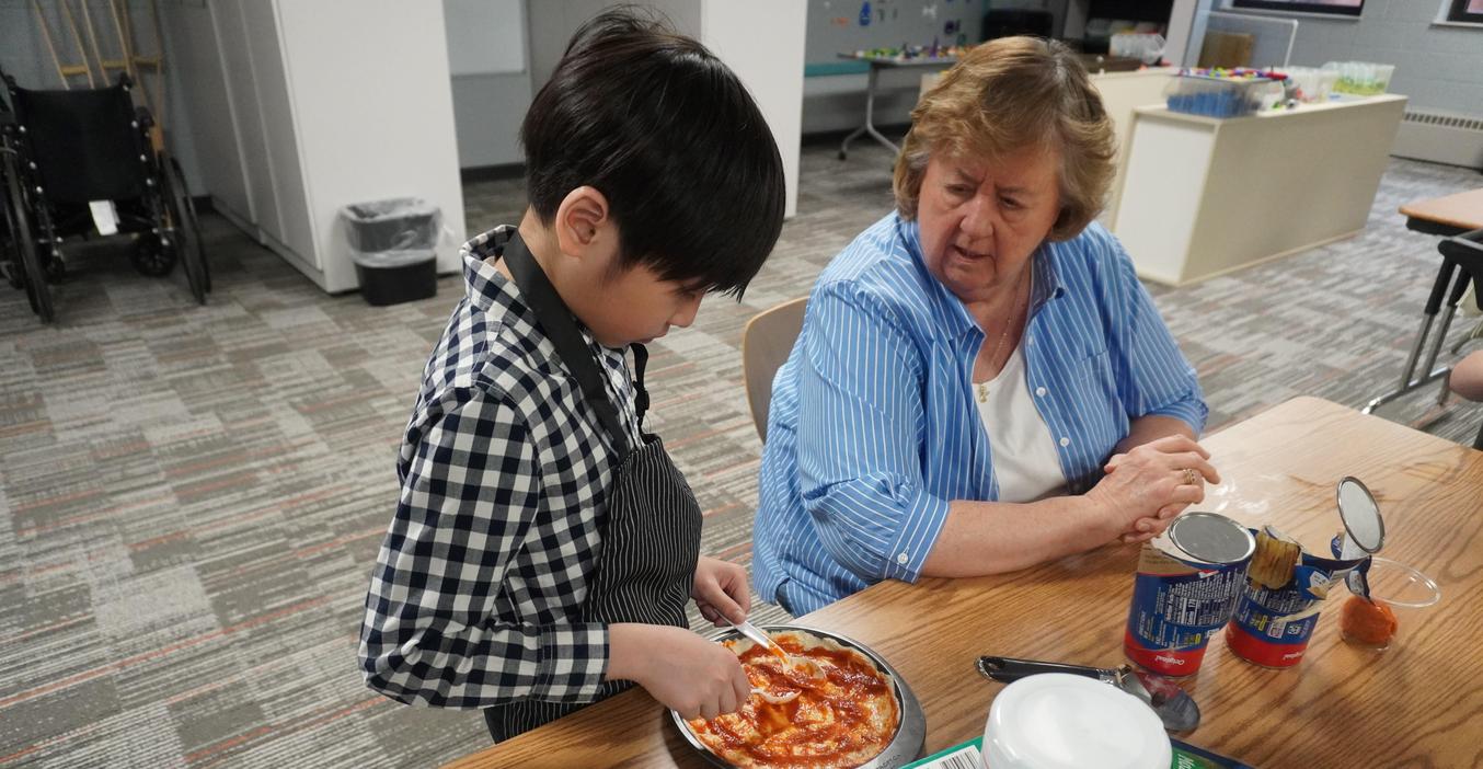 A child making a personal pizza while a volunteer grandmother watches.