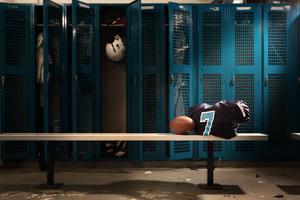 An empty sports locker room featuring a row of tall, teal mesh lockers. In the foreground, a long metal bench holds an American football propped against a folded navy blue jersey with a light blue and white number 7. One locker door in the background is open, revealing a white football helmet hanging inside.