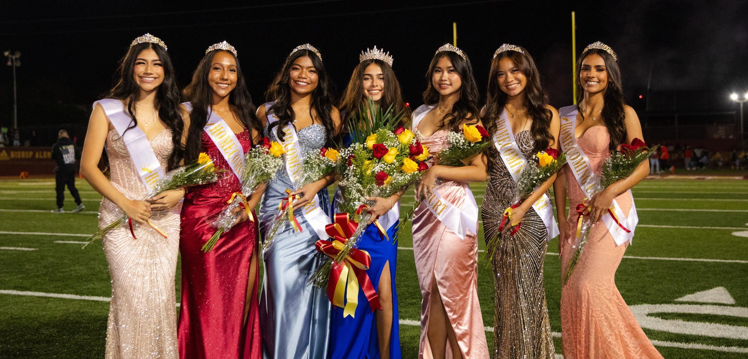 Seven crowned girls in formal dresses holding flowers on a field.