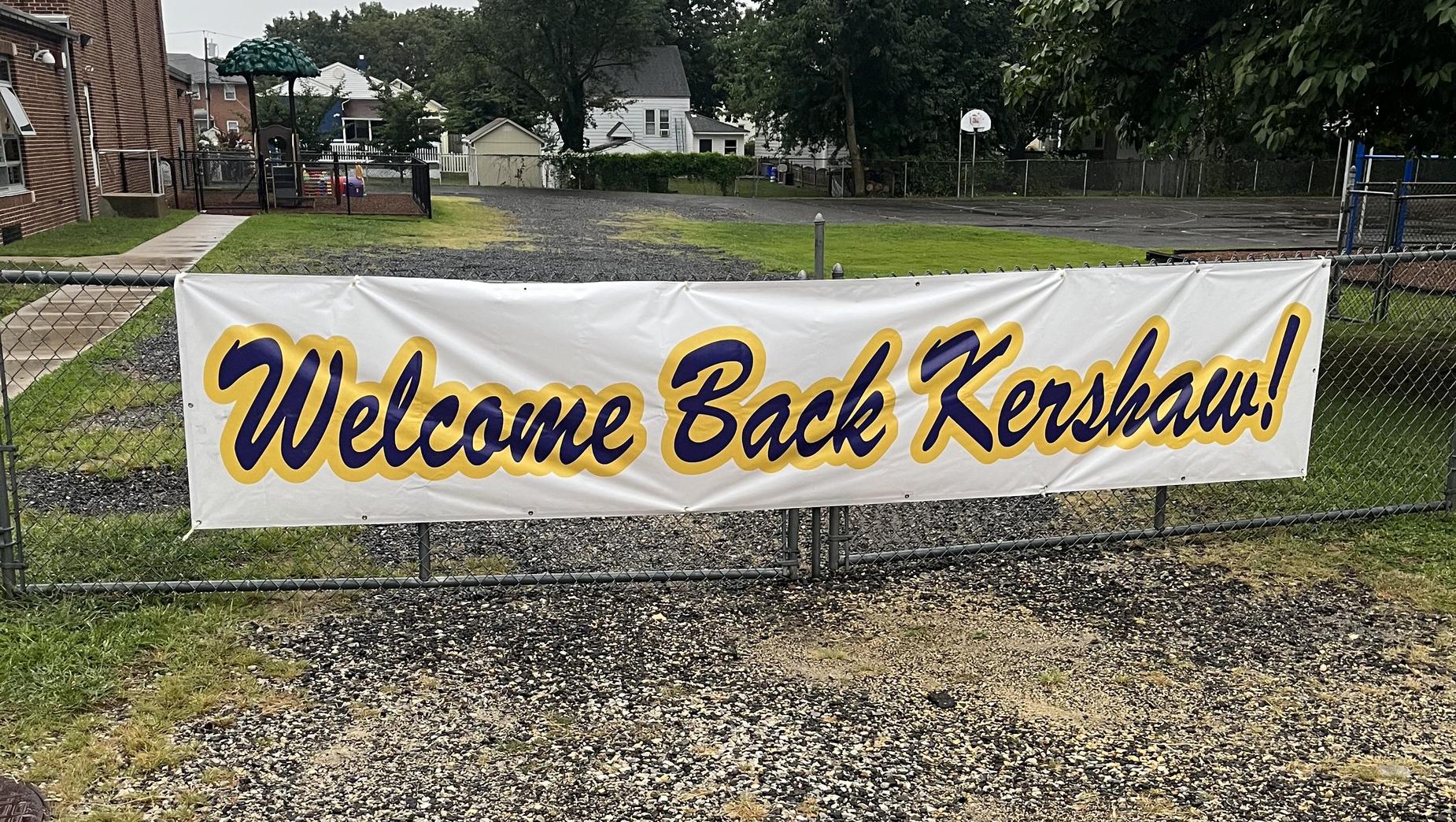 Large sign hangs on the fence outside of the school near the entrance reading