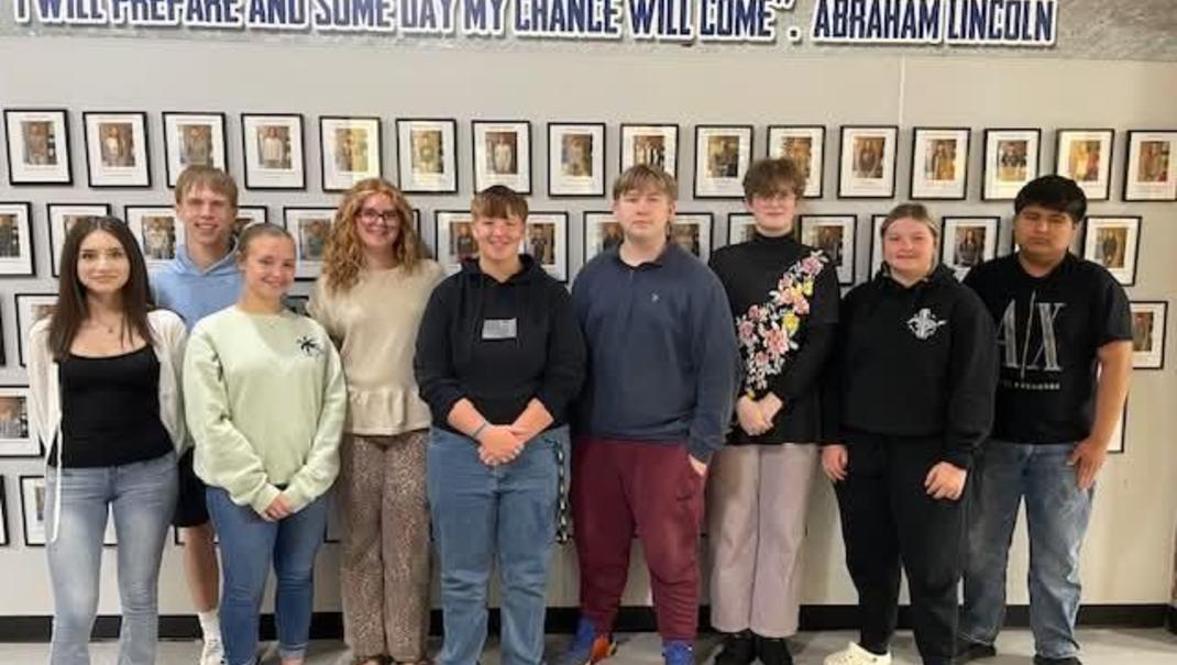 A group of students poses in front of a wall with framed pictures and a quote.