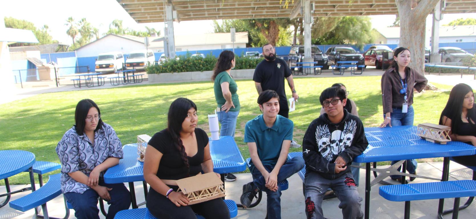 A group of people sitting at blue picnic tables in a park-like setting.
