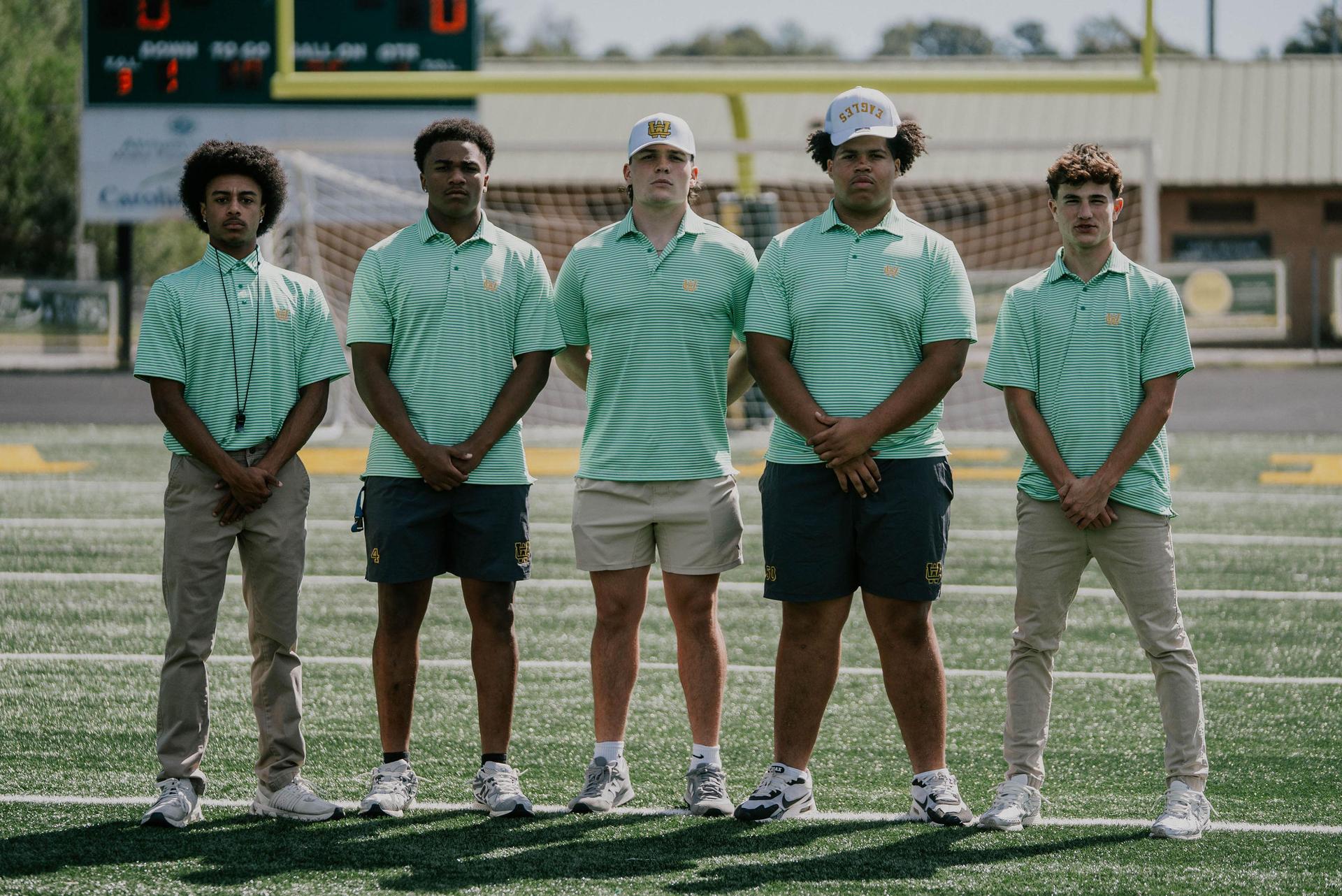 Five young men in polo shirts pose together on a football field for a team photo.