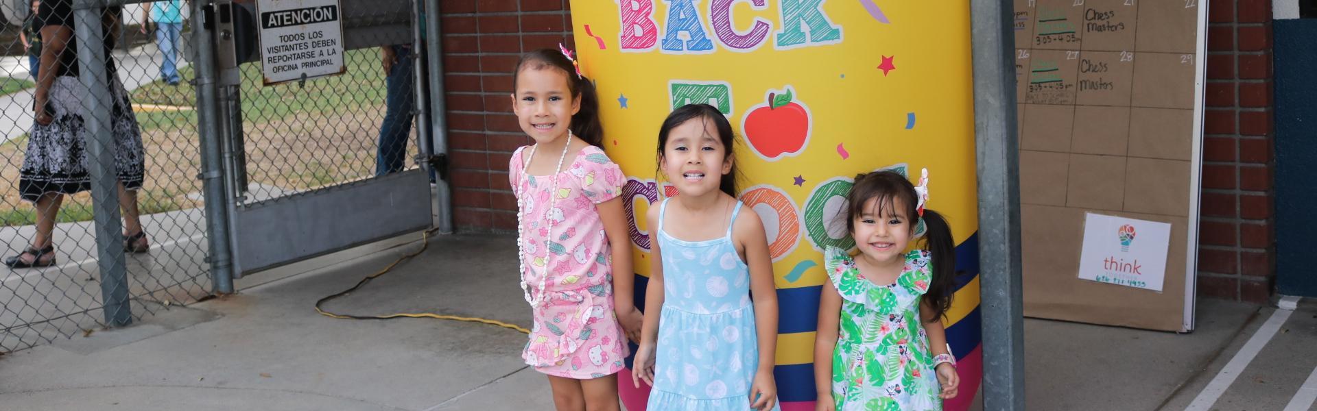 Three girls pose in front of a colorful 'Welcome Back' banner at a school entrance.