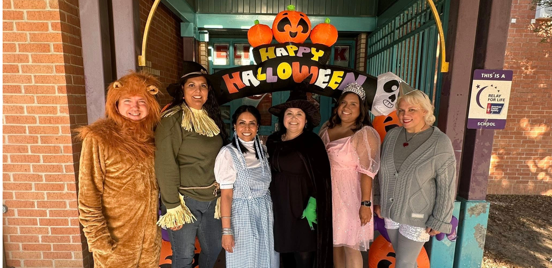 Staff dressed as Wizard of Oz characters in from of vlack, white, yellow, orange and purple Halloween sign