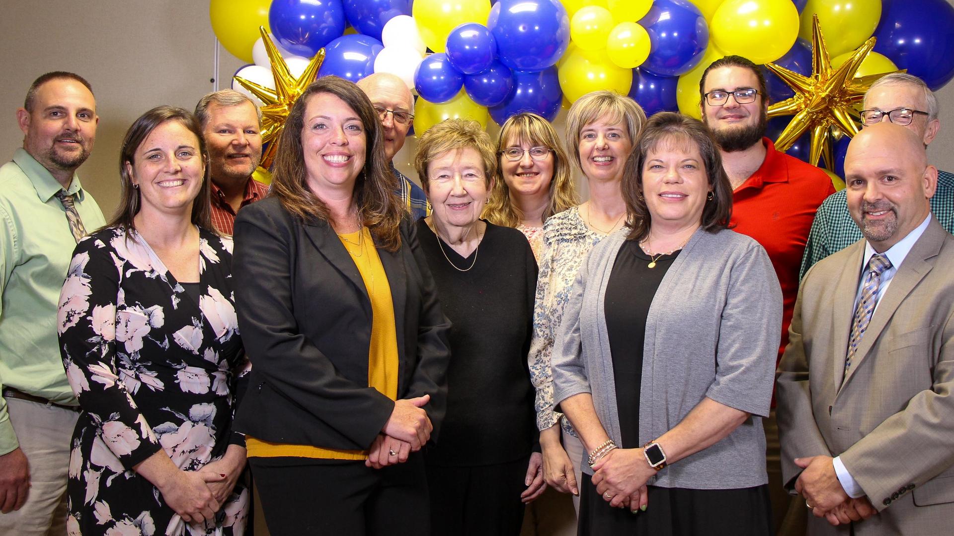 A group of IU5 Adult Education staff and volunteers smiling in front of blue and yellow balloon decorations at an Adult Education Gradution Ceremony.