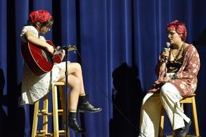 A girl plays guitar while another girl sings, both seated on stools.