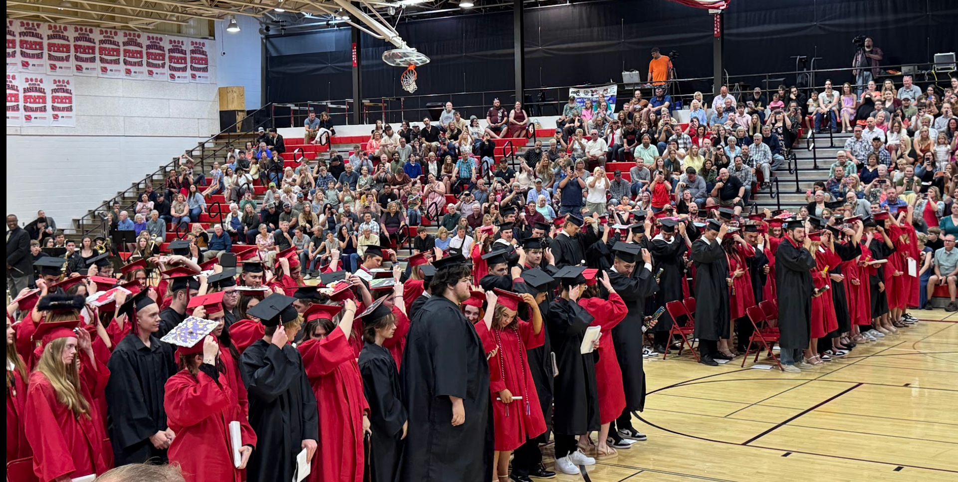Graduation ceremony with graduates in caps and gowns, filled bleachers of spectators.