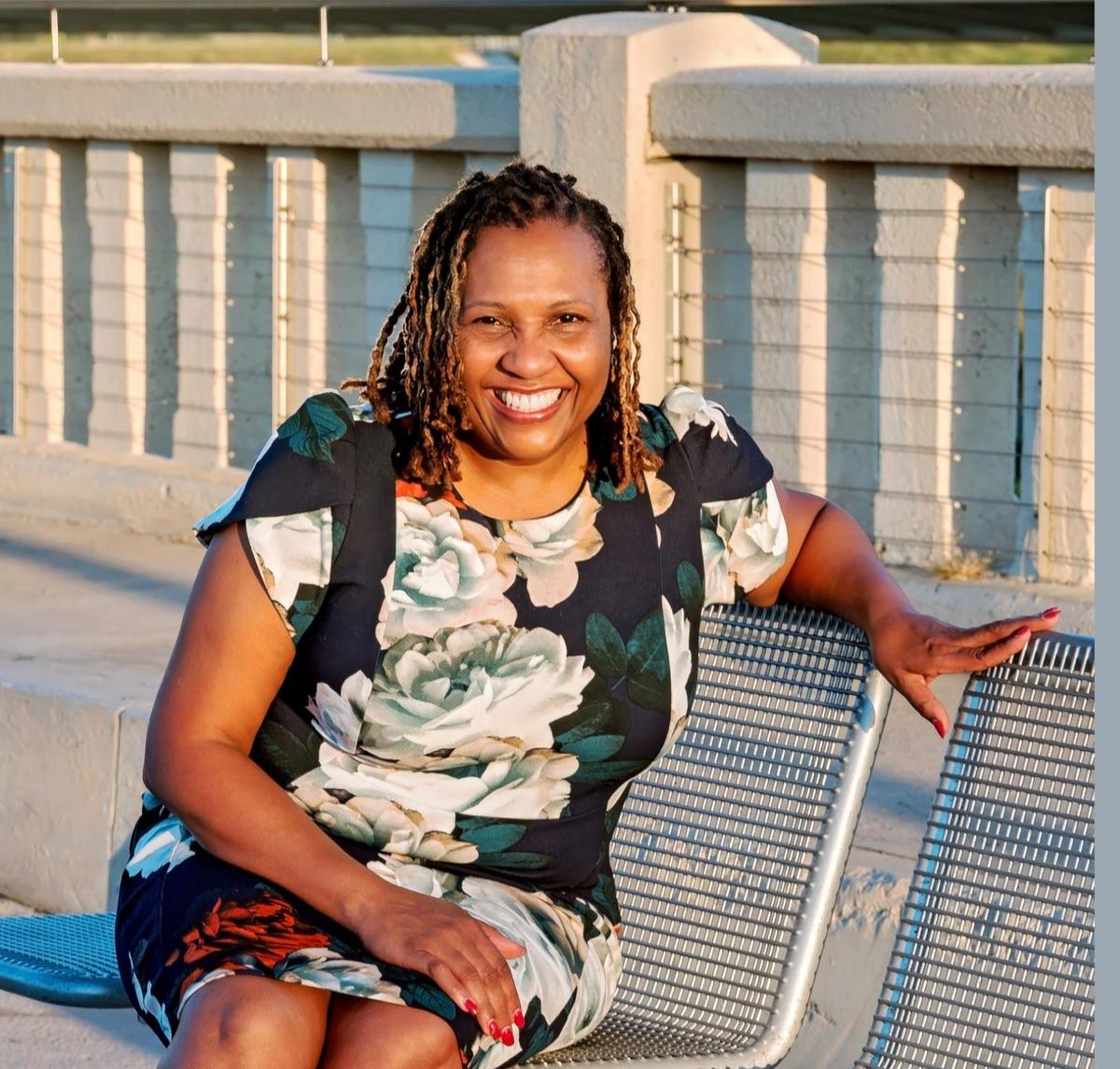 Woman with curly hair in a floral dress, sitting on a bench and smiling.