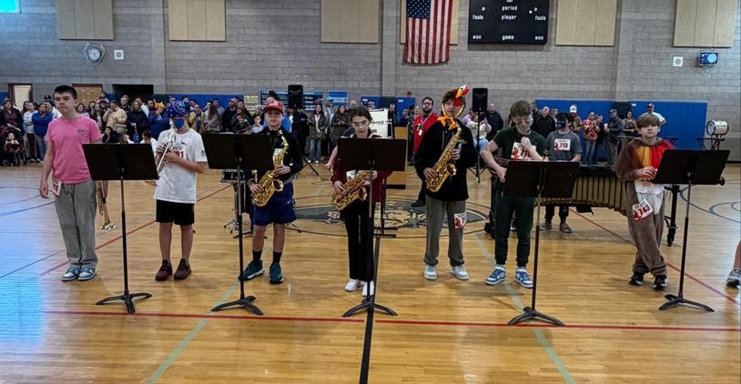 Boys playing various instruments on stage in a gym, with an audience in the background.