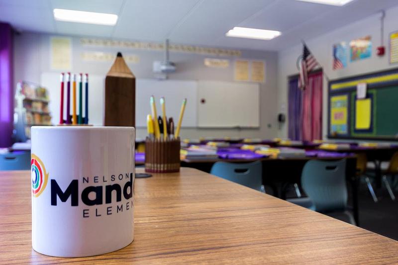 An empty classroom is pictured with a white, Mandela mug sitting on a teacher's desk, as the focus of the photo, filled with pencils,