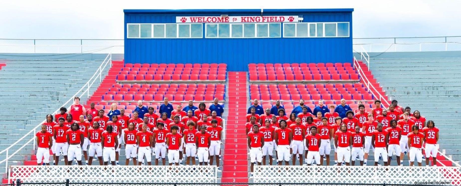 A group of high school football players in red jerseys posing in a stadium setting.