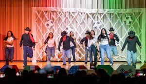 A group of students perform a lively dance on a school stage, moving forward in unison. They wear casual outfits—jeans, white or black tops—and some have cowboy hats and red bandanas. The backdrop features a lattice design decorated with soccer ball cutouts, lit with red, white, and green stage lighting. Audience members are visible in silhouette at the bottom, some holding up phones to record the performance.