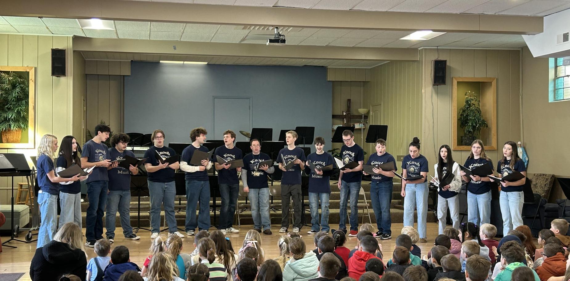 singers stand holding music while students are seated on floor in front of them
