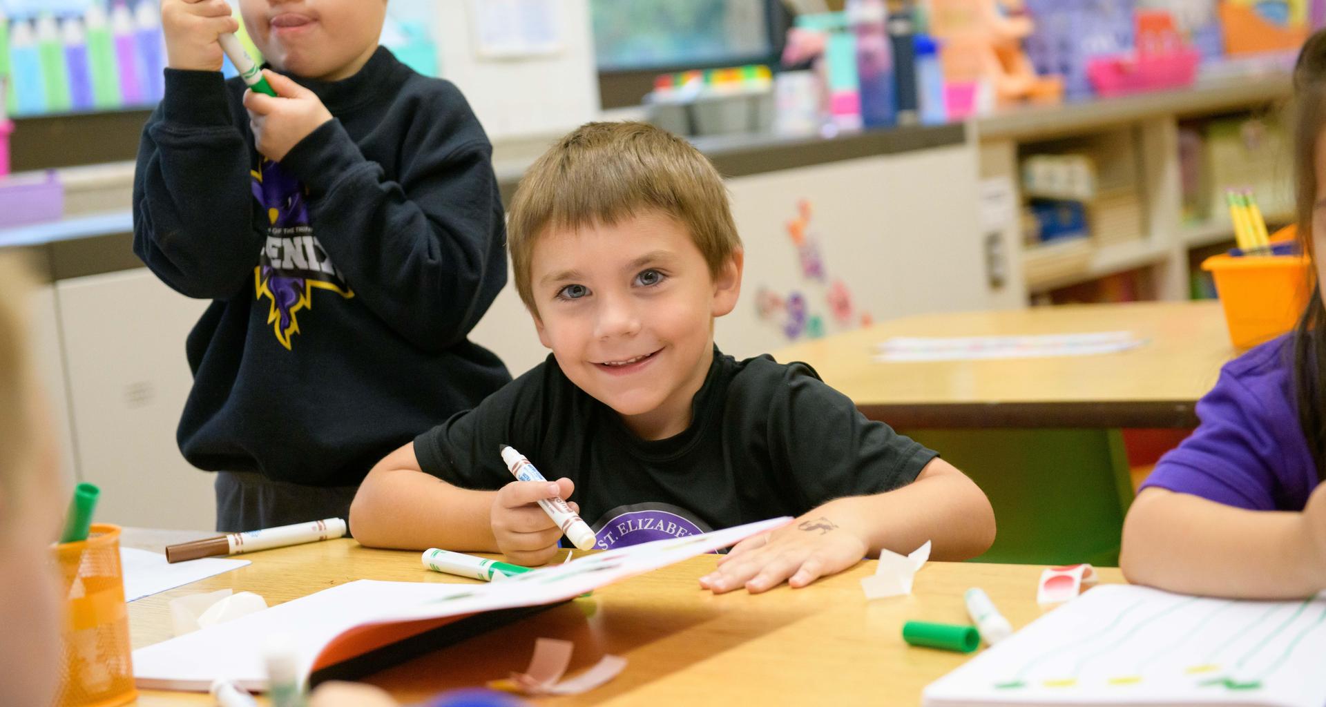 Boy coloring at a table with markers, smiling at the camera.