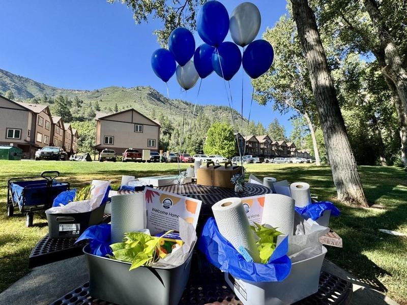 Housewarming baskets filled with gifts sit on a picnic table, with blue and white balloons and apartment buildings in the background.