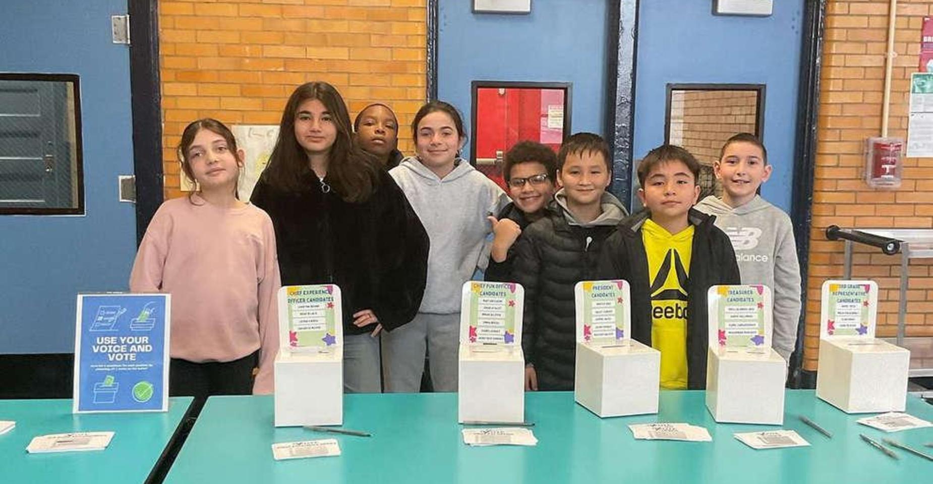 Group of children standing at a voting booth with boxes and signs.
