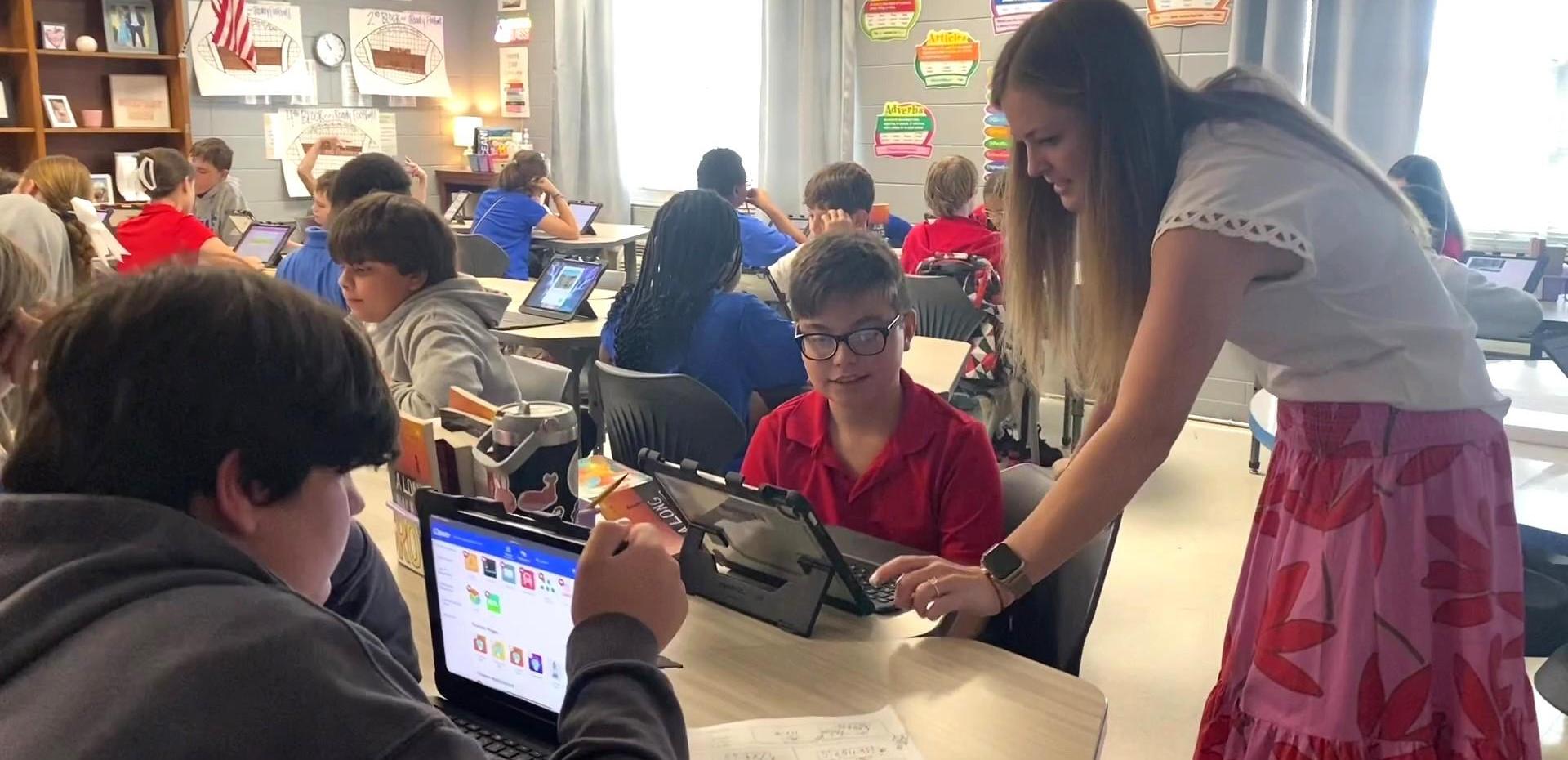 Teacher assisting a student with technology in a lively classroom filled with engaged learners.