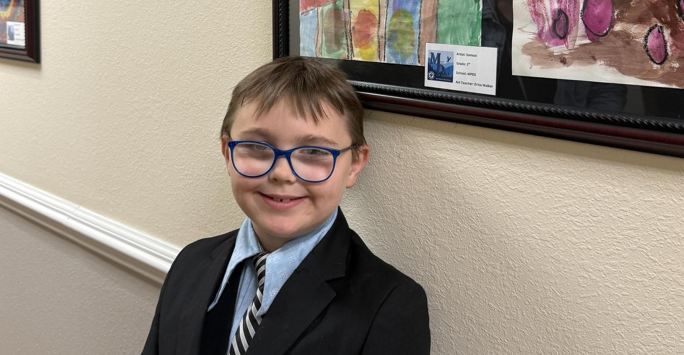 A smiling boy wearing glasses stands next to colorful artwork in a gallery.