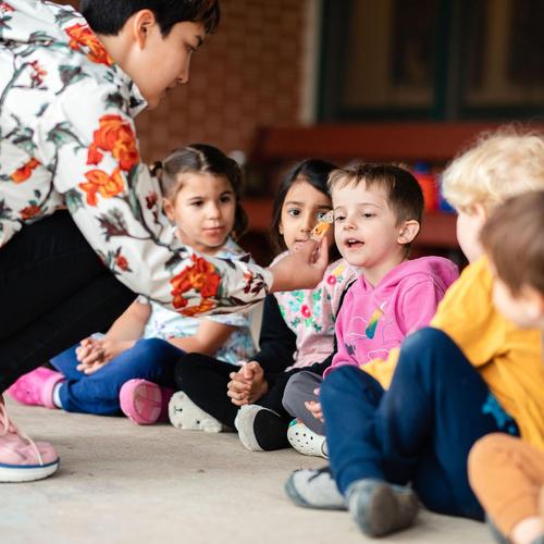 A caregiver feeding a child while several kids watch eagerly.