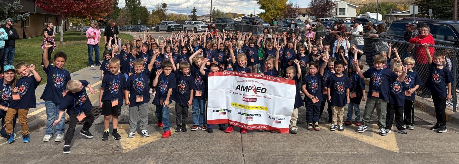 Large group of children wearing matching shirts, holding hands in the air at an outdoor event.