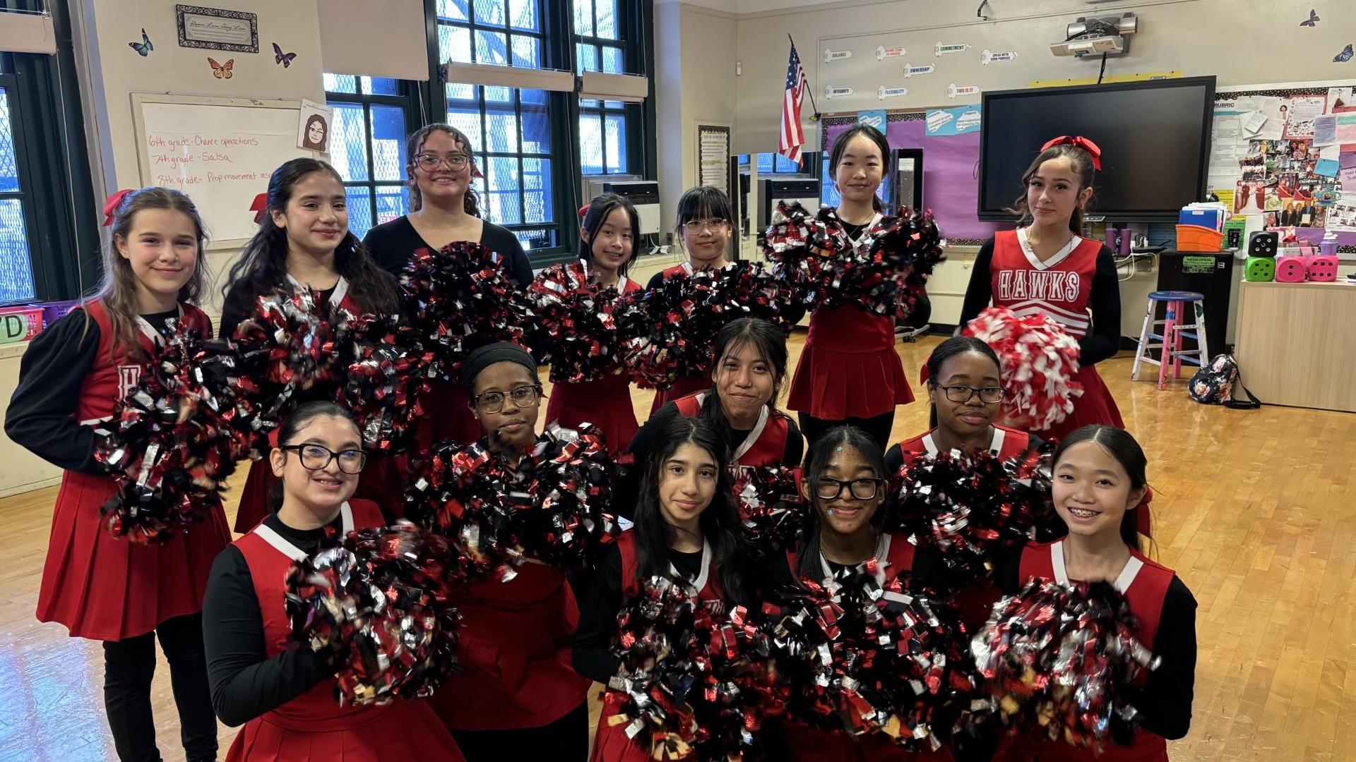 A group of cheerleaders in red uniforms holding pom-poms, smiling in a classroom.