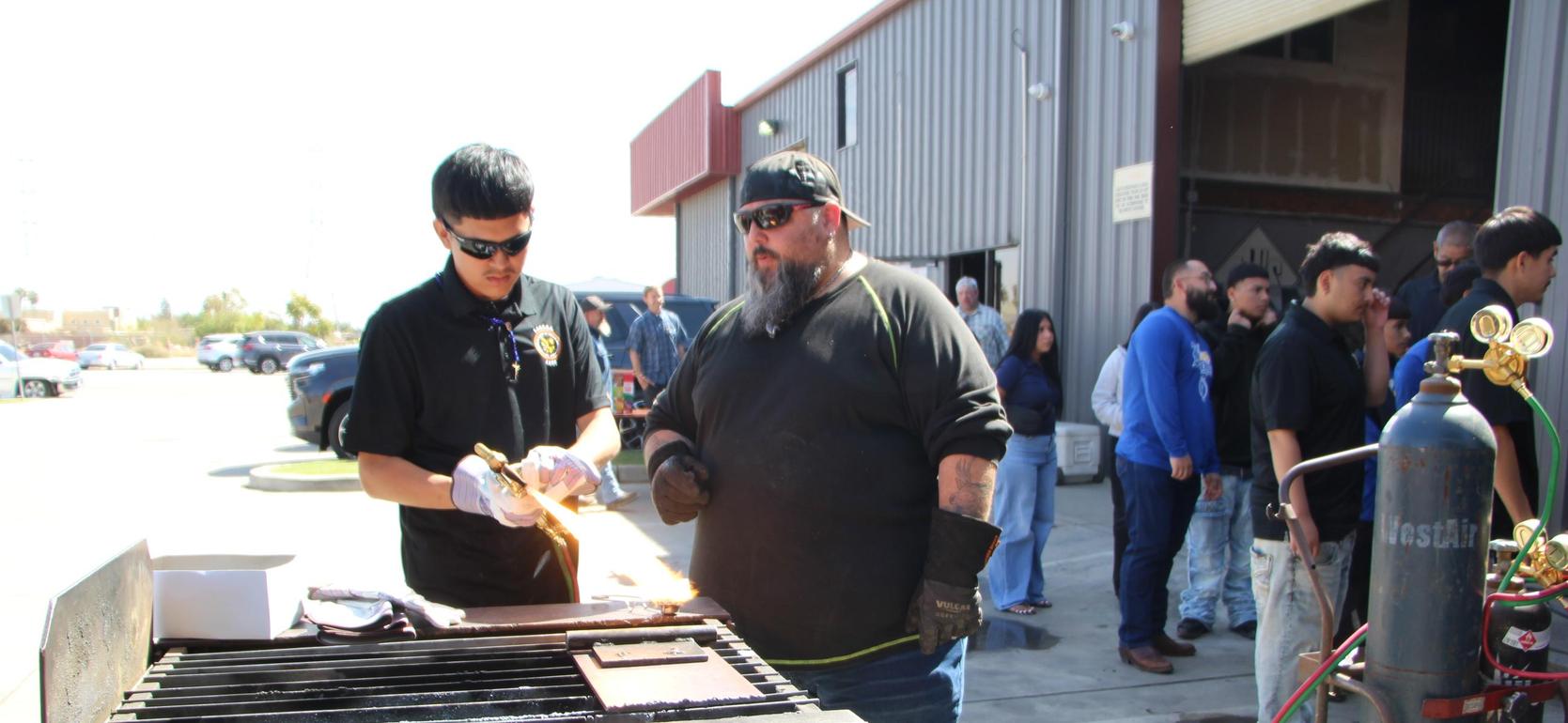 Two men in a workshop; one using a torch on metal while others observe.