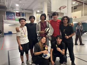 Group of seven individuals with trophies and sports equipment in a gym setting.