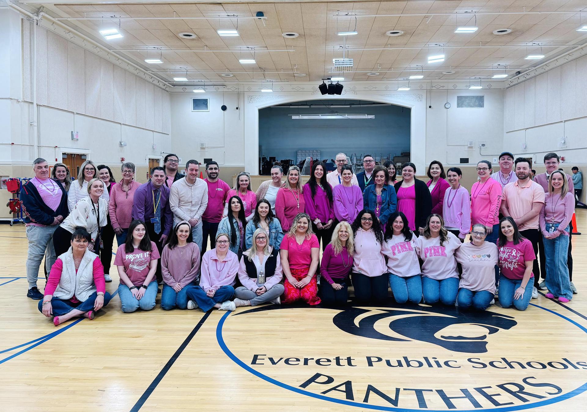 Group of people in pink attire smiling for a photo in a gymnasium.