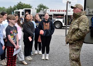 A military personnel addresses a group of students on an outdoor training site.