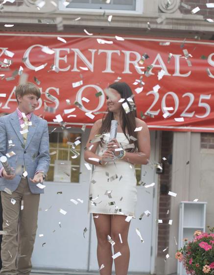 Confetti falls as two students celebrate graduation in front of a school banner.