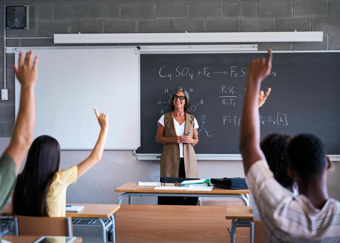 Teacher in a classroom with students raising hands for questions.
