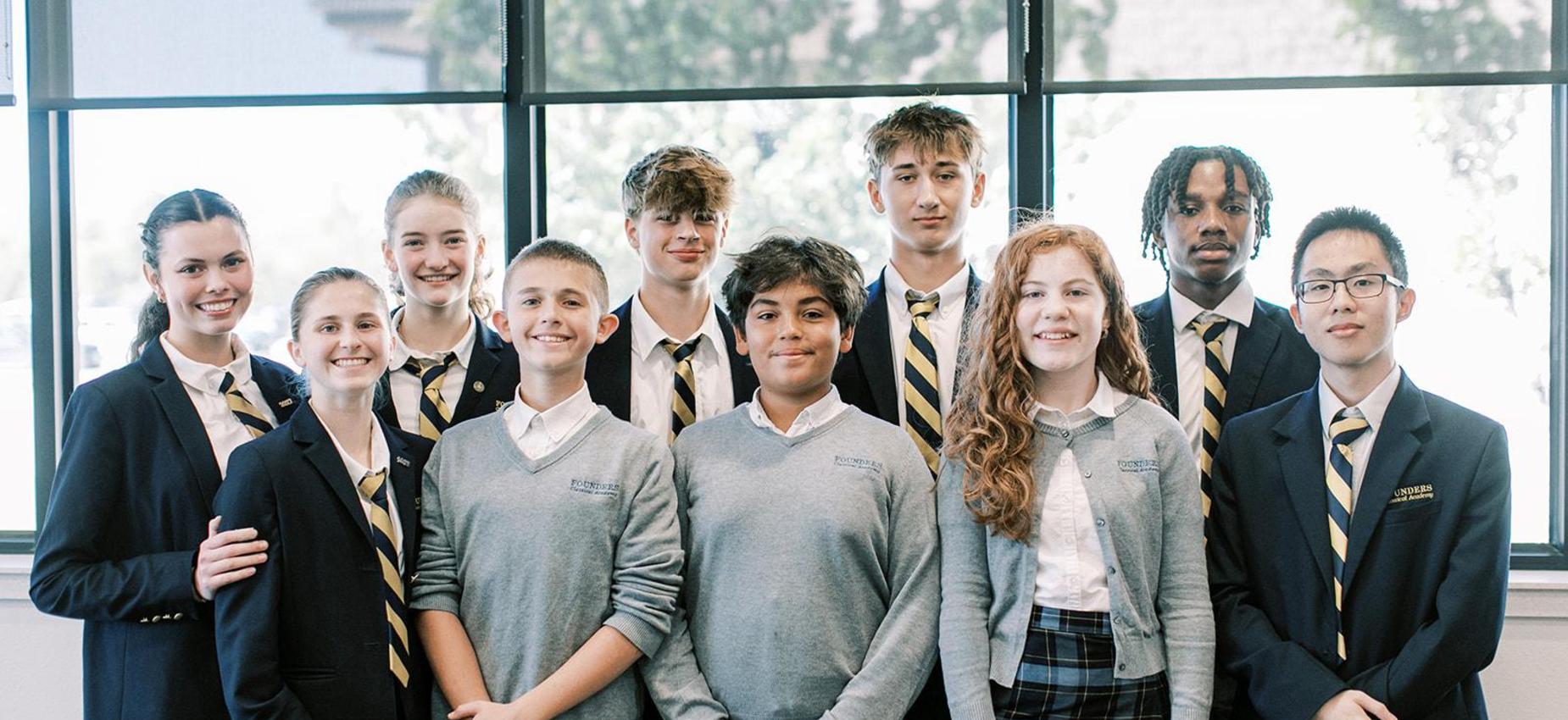 Group of students in blazers and sweaters stands together by large windows