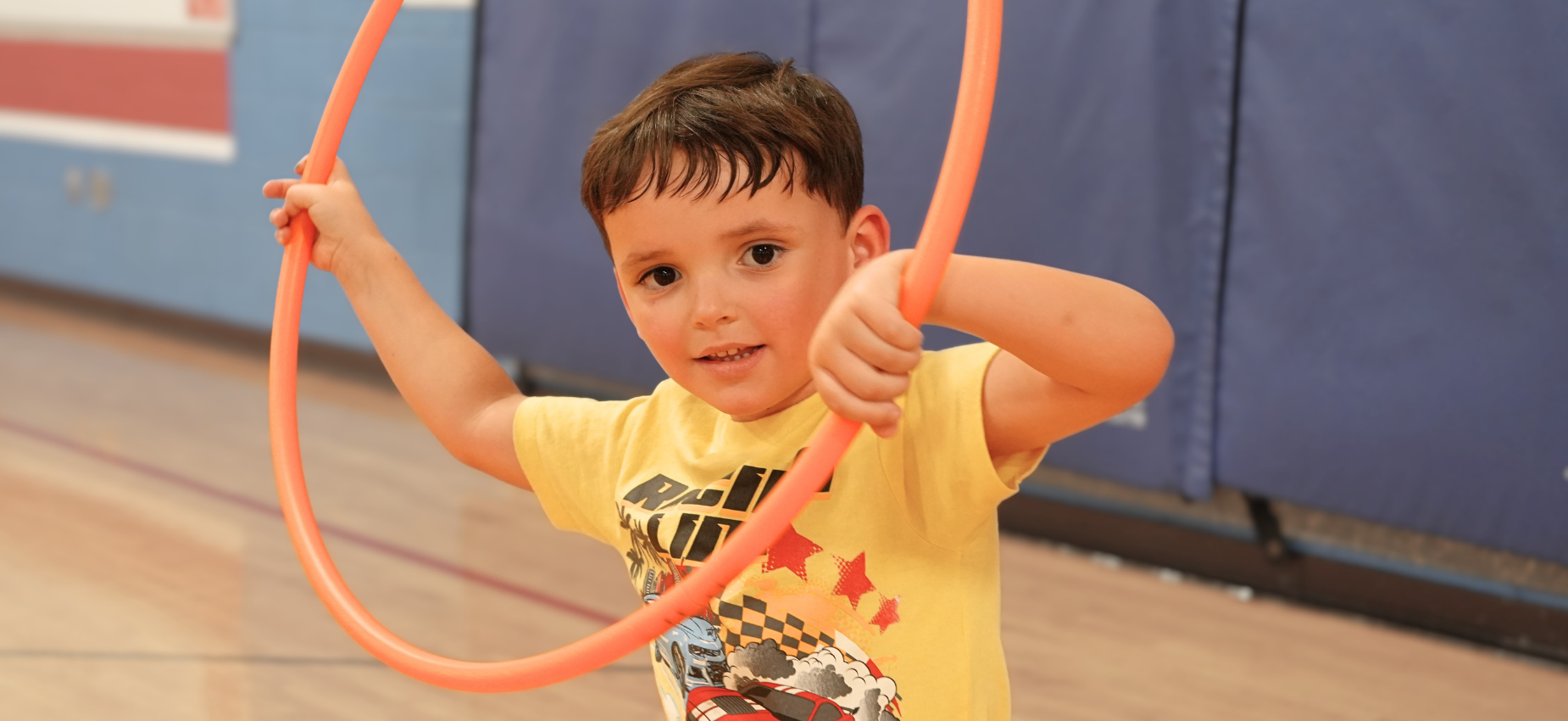 Child in a yellow shirt playing with an orange hula hoop in a gym.