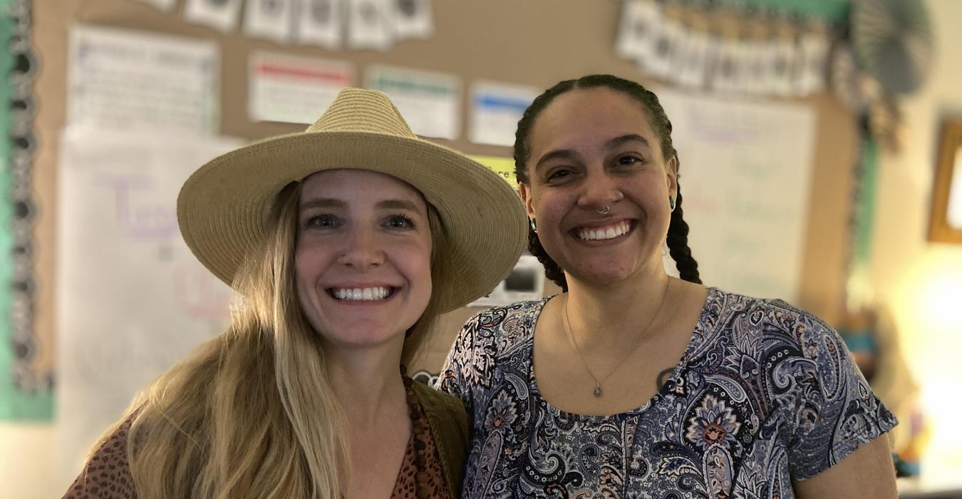 Two smiling women in a classroom, one wearing a wide-brimmed hat.