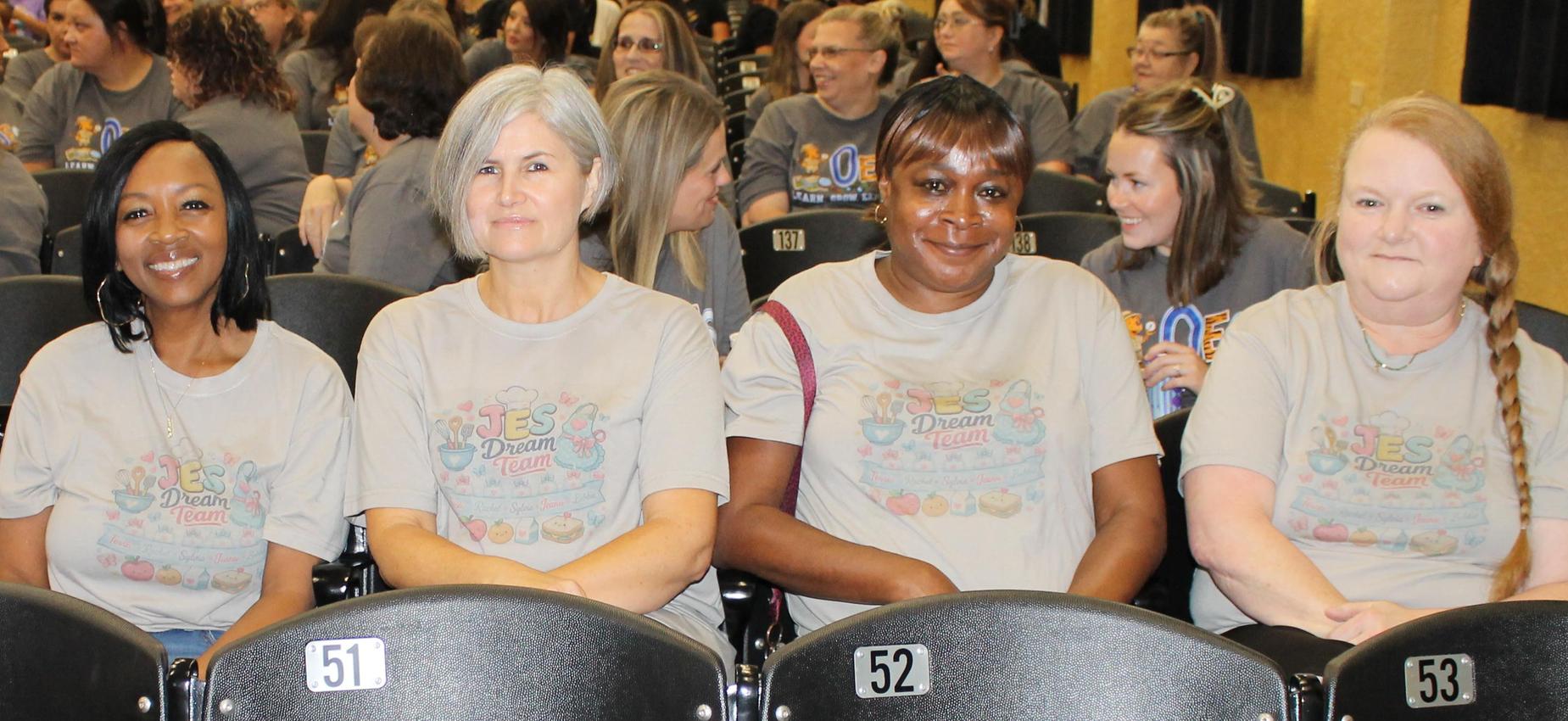 Five women in matching t-shirts seated in a row of chairs.