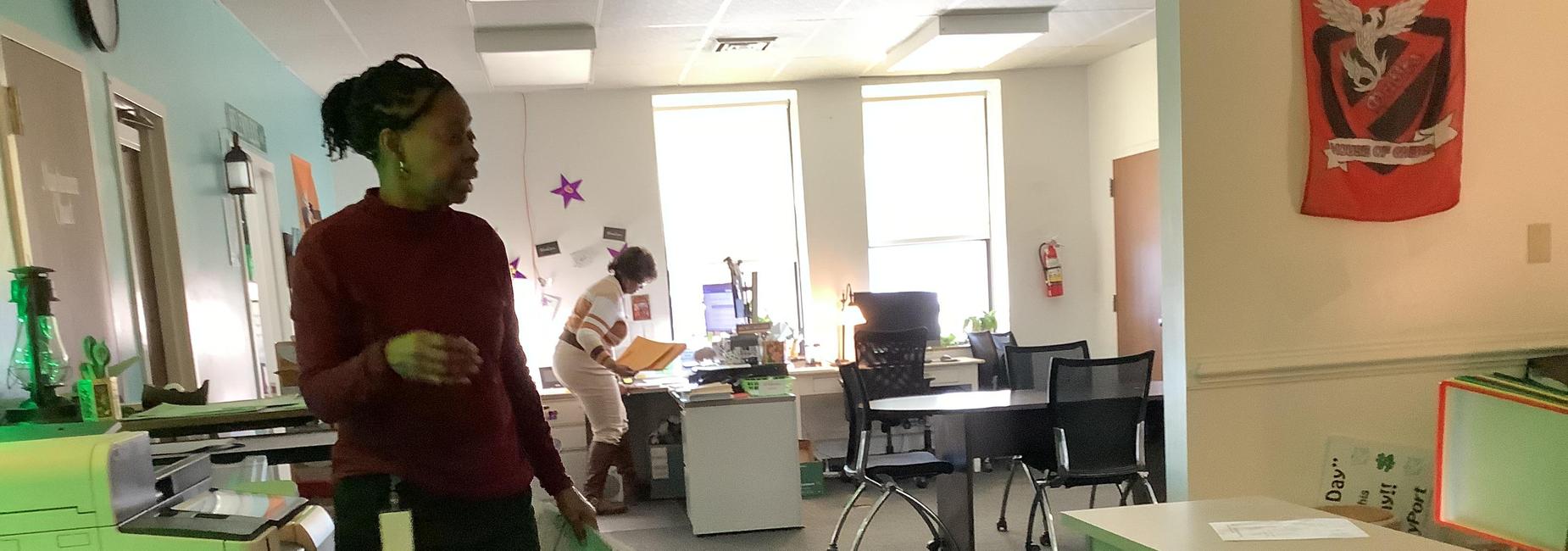 One person at a printer and another at a desk in a busy office space.