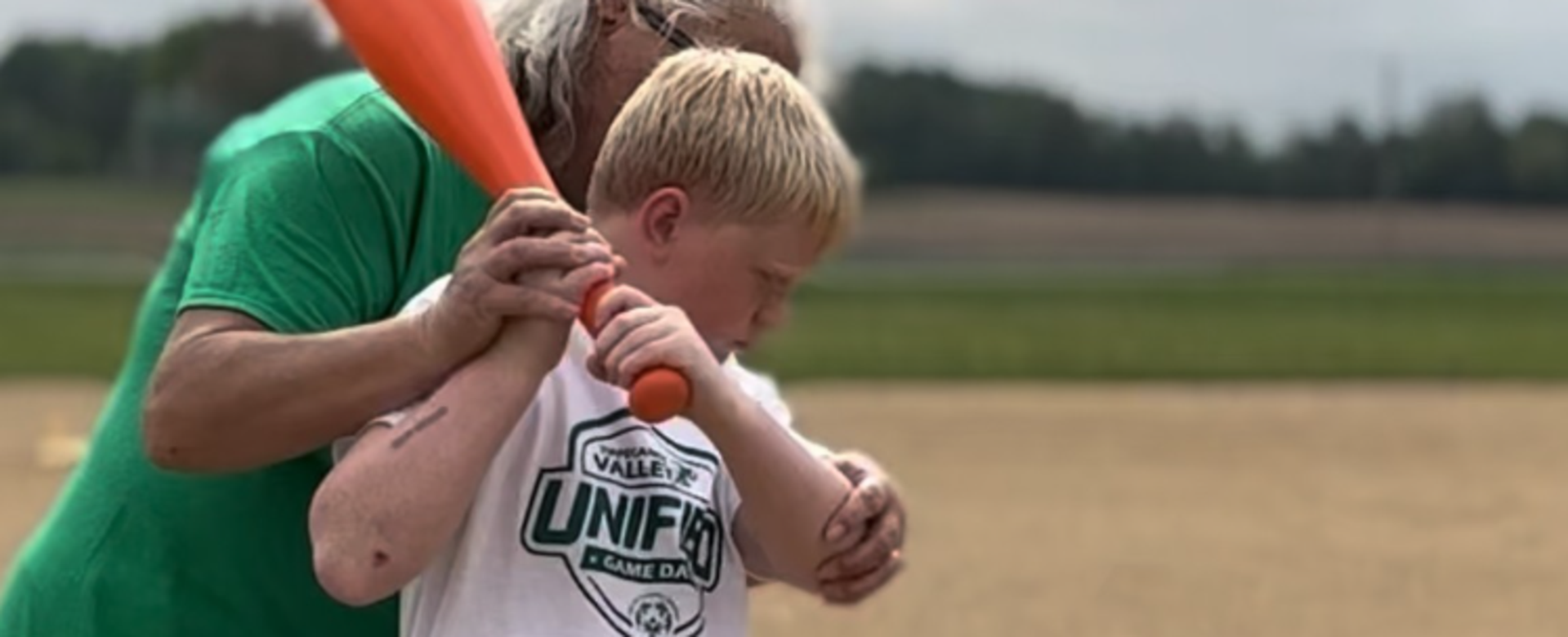 Child learning to bat with assistance from an adult on a sports field.