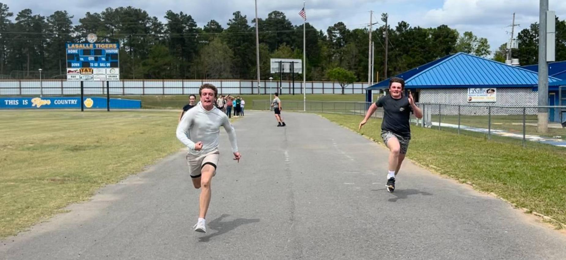 Two boys sprinting down a track with a sports field and scoreboard in the background.